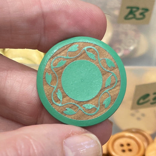 Hand holding a decorative button in front of a container filled with various buttons.