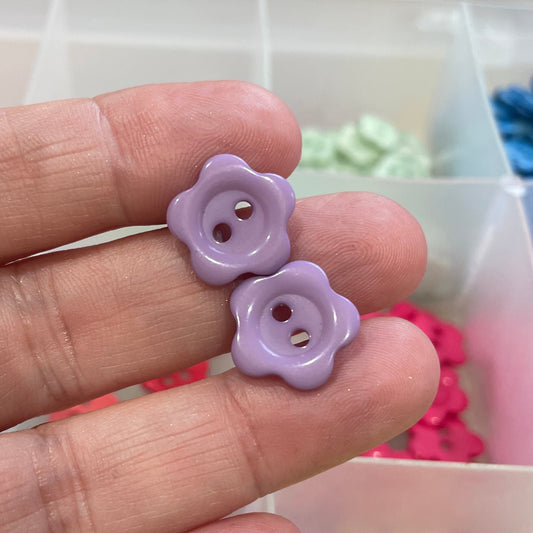 Hand holding a purple flower-shaped button with a container of colorful buttons in the background.
