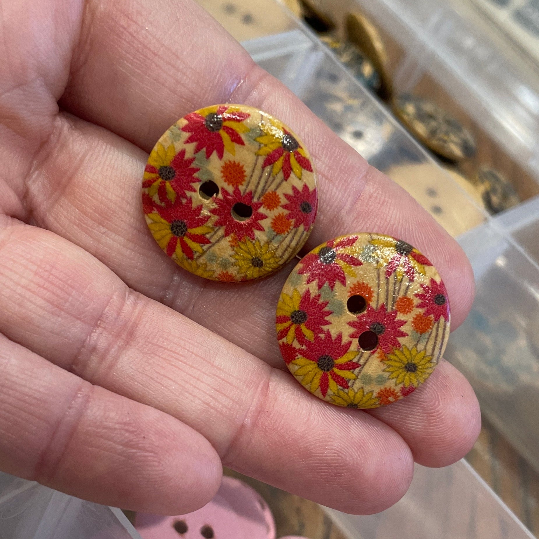 Hand holding two floral-patterned buttons with a container of assorted buttons in the background.