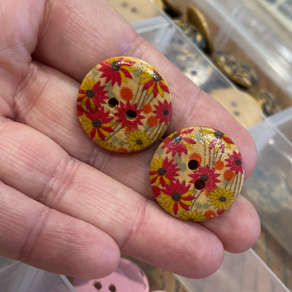 Hand holding two floral-patterned buttons with a container of assorted buttons in the background.