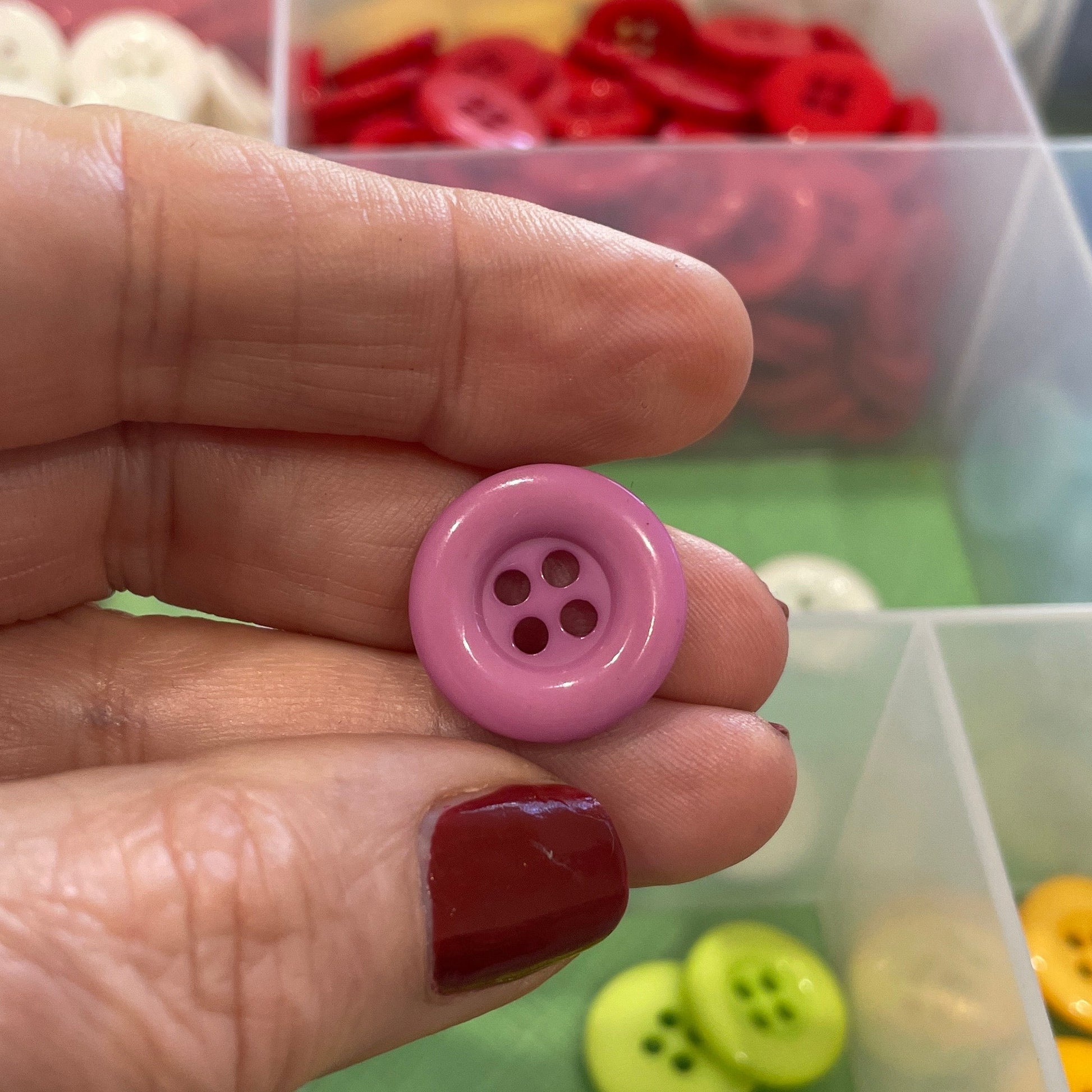 Hand holding a pink button with a container of colorful buttons in the background