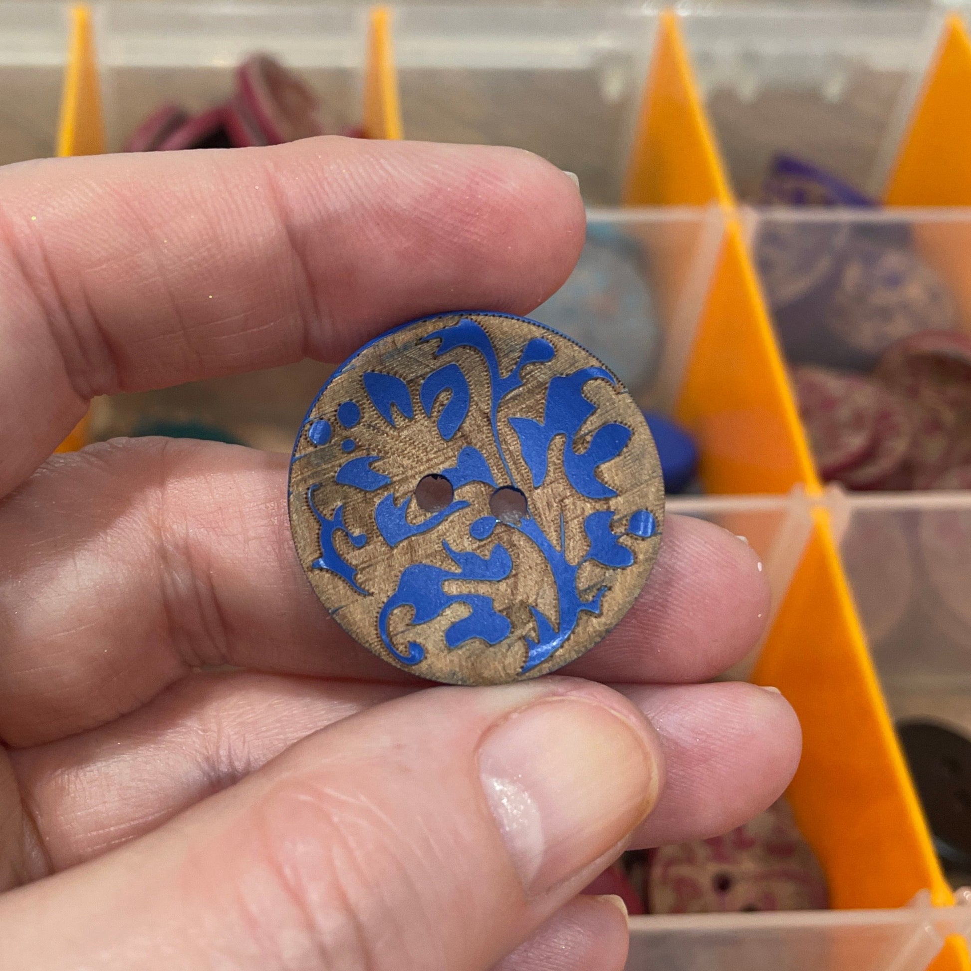 Hand holding a decorative button with intricate design in front of a button organizer.