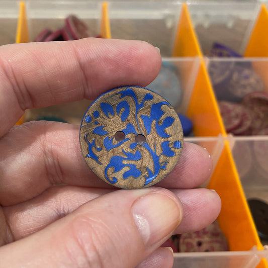 Hand holding a decorative button with intricate design in front of a button organizer.