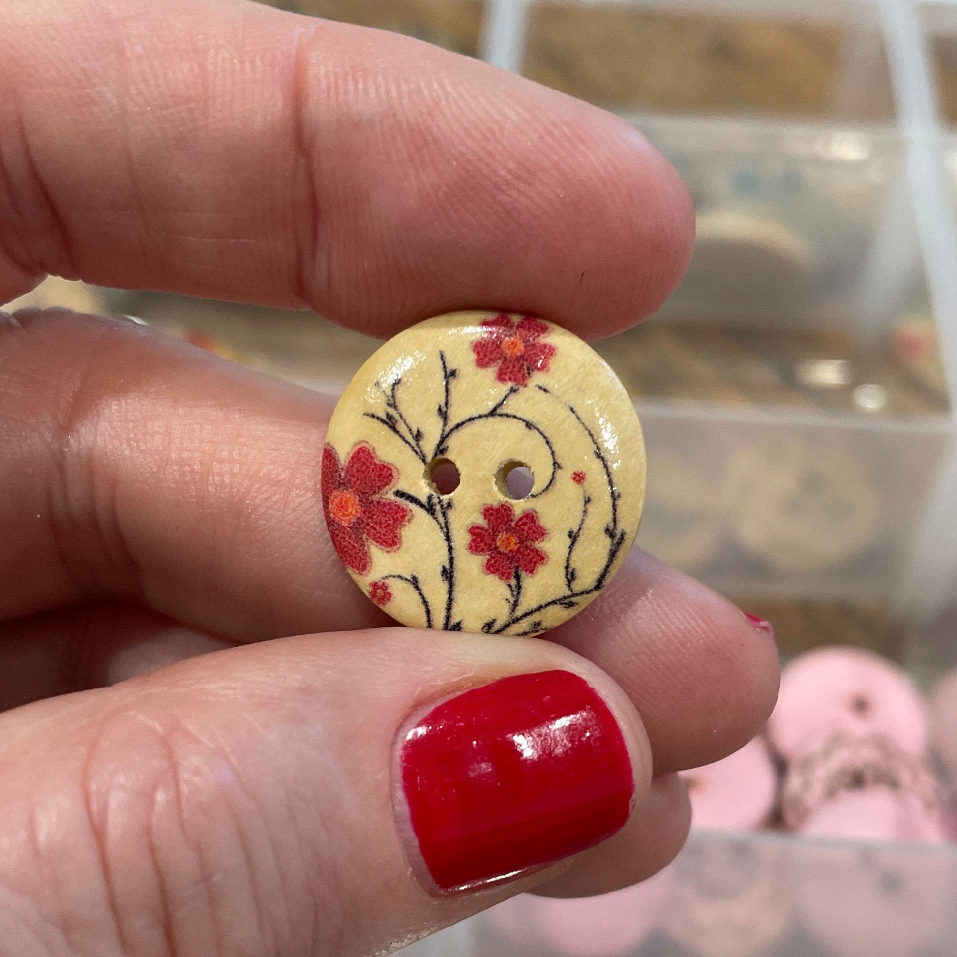 Hand holding a decorative button with a floral design in front of a container of buttons.