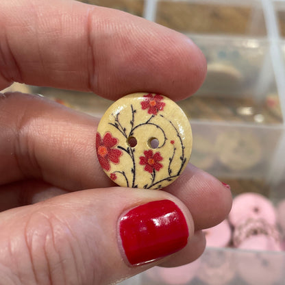 Hand holding a decorative button with a floral design in front of a container of buttons.