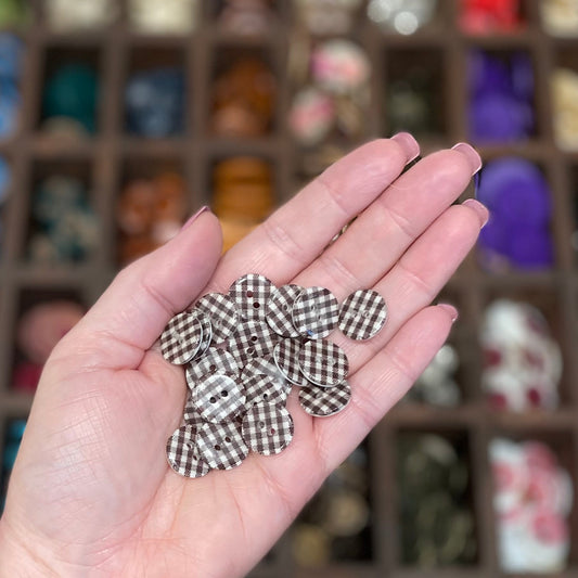 A person's hand holding a brown and white gingham resin button with a box of various buttons in the background.