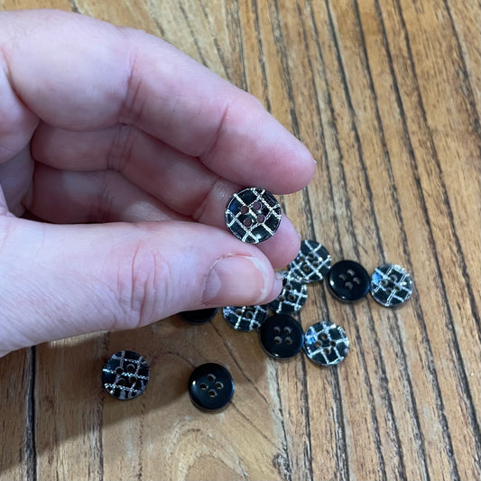 Hand holding a black and silver button on a wooden surface with more buttons scattered around.