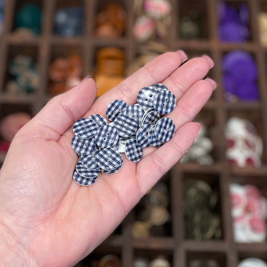 A person's hand holding several navy blue and white gingham resin buttons with a blurred background of various craft materials.