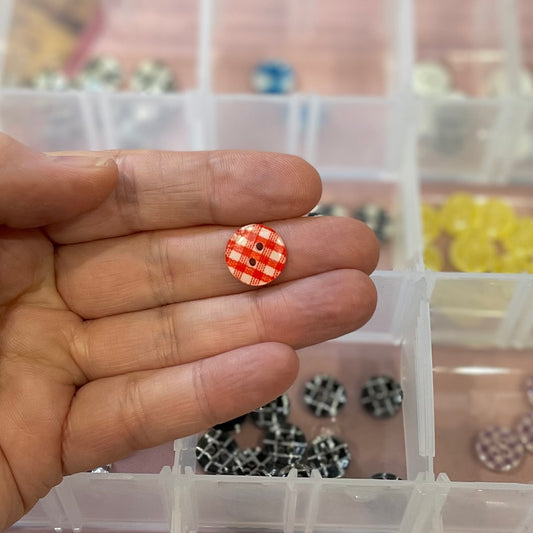 Hand holding a small orange and white checkered button with a blurred background of a container with more buttons.