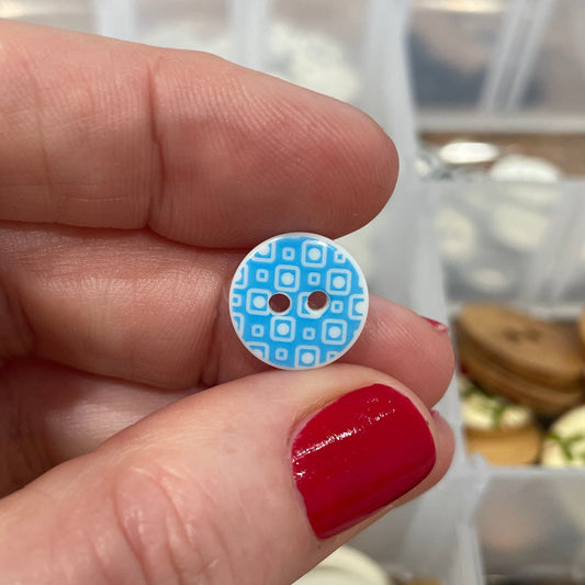 Hand holding a blue button with a pattern, with a container of buttons in the background.