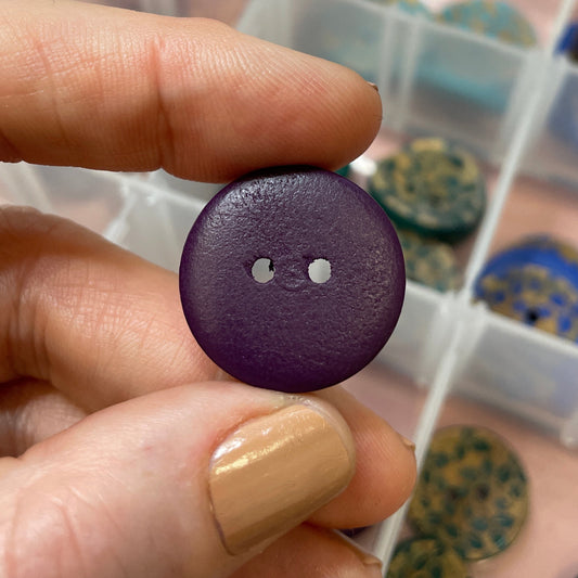 Hand holding a purple button with a container of buttons in the background