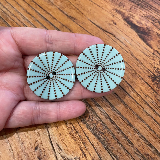 Two circular buttons with a brown dot pattern held between fingers on a wooden surface.