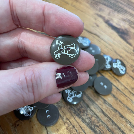 Hand holding a button with a scooter design on a wooden surface with more buttons.