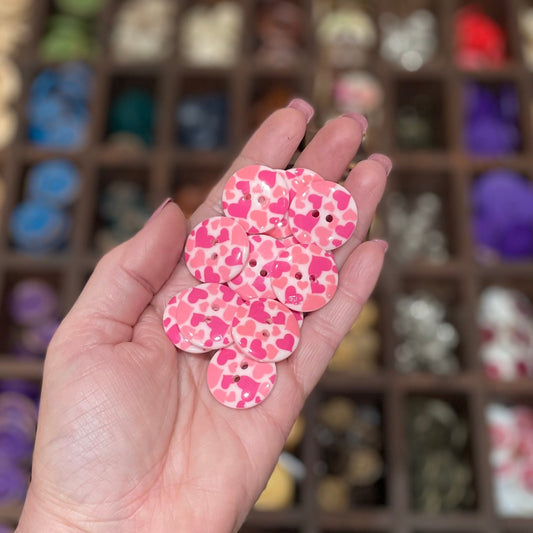 A hand holding a few pale pink resin buttons with a heart pattern and two holes each.