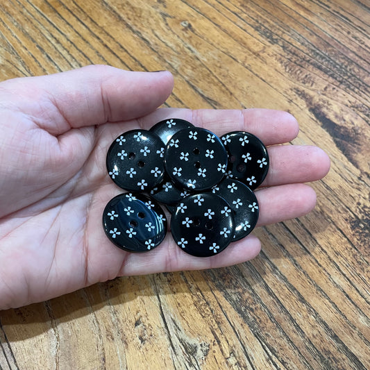 Hand holding black buttons with white floral patterns on a wooden surface