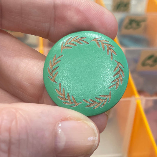 Green decorative button held by a hand with a blurred background of colorful buttons in a case.