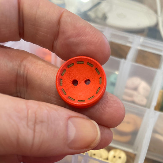 Hand holding an orange button with a container of colorful buttons in the background.