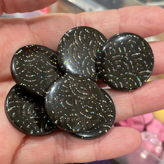 Hand holding black buttons with glittery specks, with a container of colorful buttons in the background.