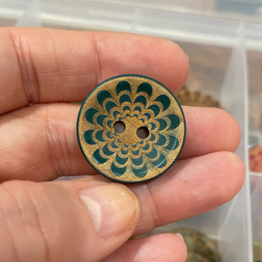 Hand holding a decorative wooden button with a detailed design, set against a background of similar buttons in a box.