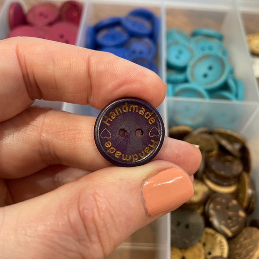 Hand holding a button with 'Handmade' text in front of a container of assorted buttons.