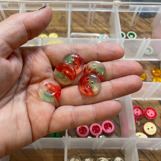 Hand holding small decorative buttons with a clear container of more buttons in the background.