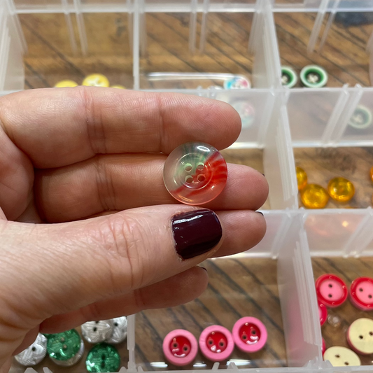 Hand holding a small glass button with a decorative design in front of a container of assorted buttons.