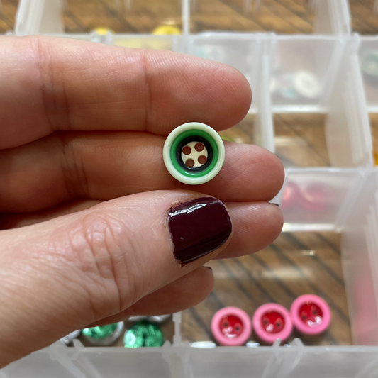 Hand holding a small green and white button with a box of buttons in the background