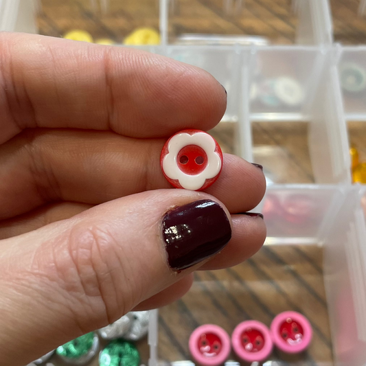 Hand holding a small red and white button with a container of buttons in the background