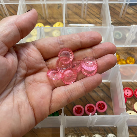 Hand holding pink buttons with a container of various buttons in the background.