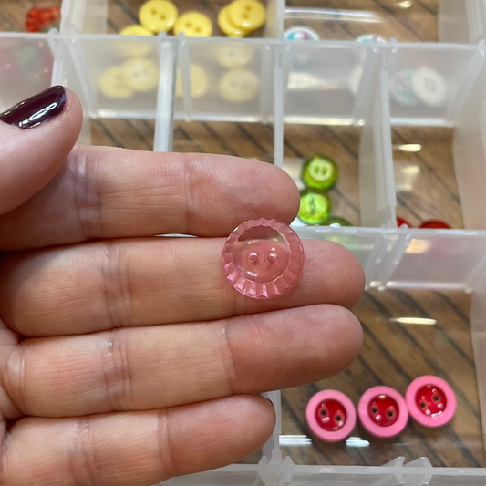 Hand holding a pink button with a clear container of various buttons in the background