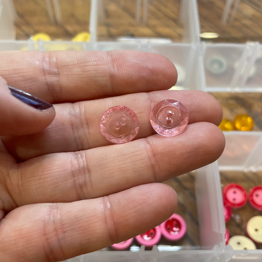 Hand holding two small pink buttons with a container of buttons in the background