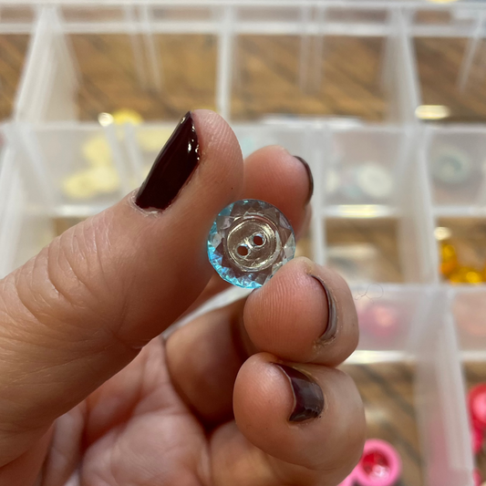 Hand holding a clear button with a metal core against a background of a clear container with buttons.