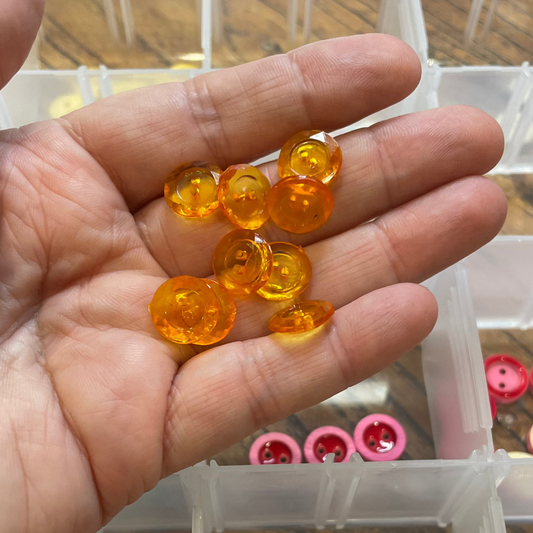 Hand holding small orange buttons above a clear container with various buttons.