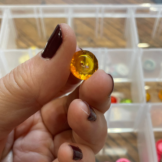 Hand holding a small orange button with a clear container of assorted buttons in the background.