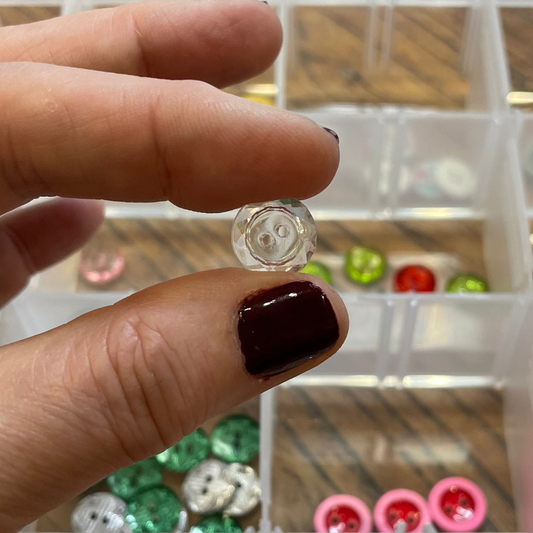 Hand holding a clear button above a container of assorted buttons on a wooden surface