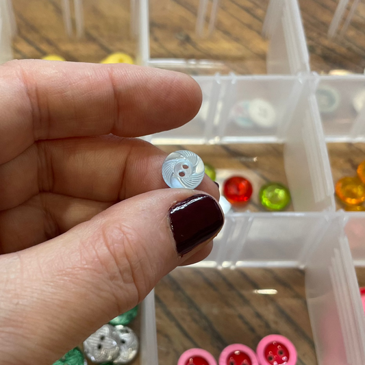 Hand holding a button above a container of various buttons on a wooden surface