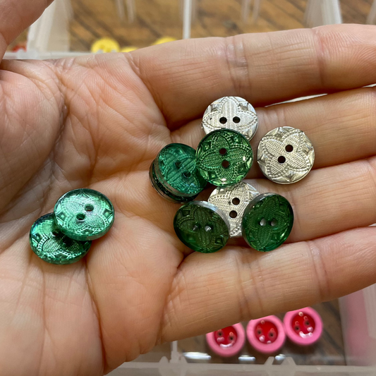 Hand holding green and silver buttons with a container of buttons in the background.