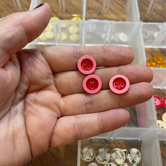 Hand holding three pink buttons with a clear container of buttons in the background.