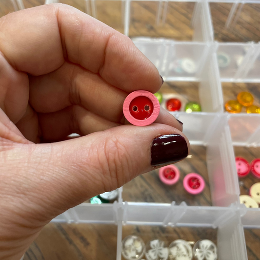 Hand holding a small pink button with a clear container of buttons in the background