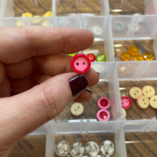 Hand holding a red button with a clear container of assorted buttons in the background