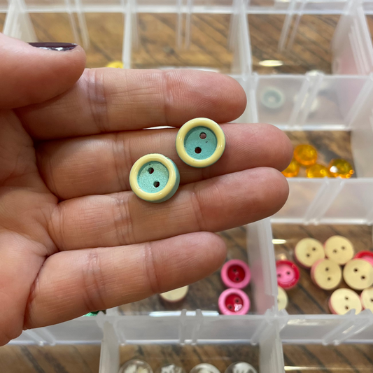 Hand holding two small buttons with a clear container of assorted buttons in the background.