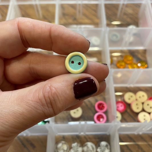 Hand holding a small button with a green face and black eyes, against a background of a button organizer.