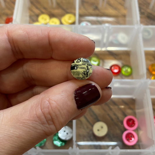 Hand holding a small button with a clear container of assorted buttons in the background.