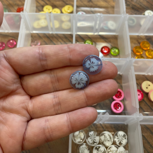Hand holding two small buttons with a clear container of buttons in the background.
