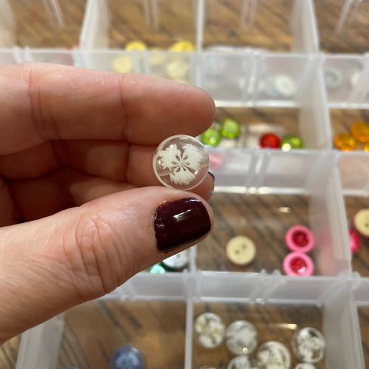Close-up of a hand holding a decorative button with a clear background showing more buttons in a container.