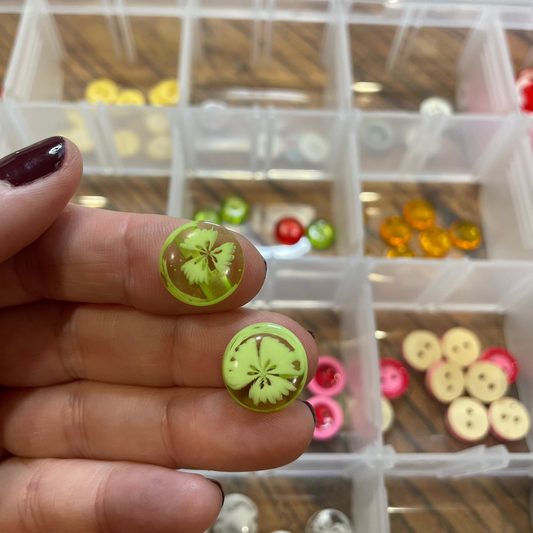 Green buttons  held between fingers with a container of buttons in the background.