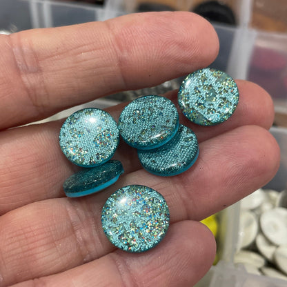 Hand holding blue glittery buttons with a blurred background of button containers.