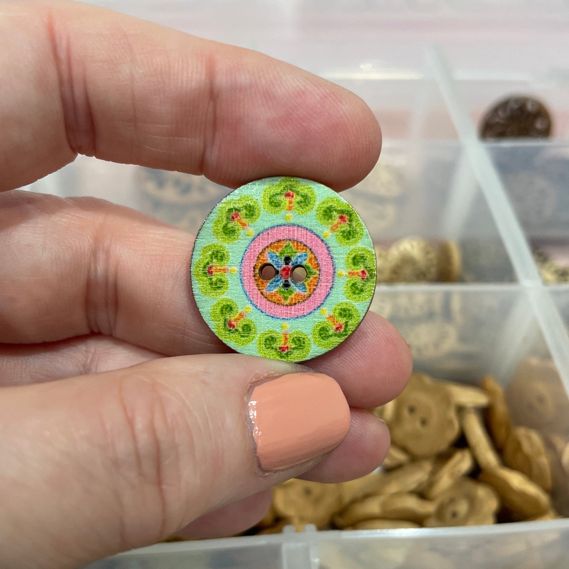 Hand holding a decorative button with a colorful design, surrounded by other buttons in a container.