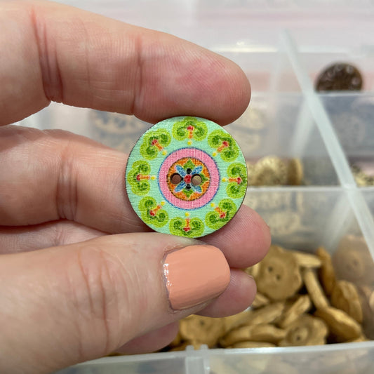 Hand holding a decorative button with a colorful design, surrounded by other buttons in a container.