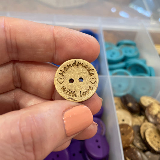 button with 'Handmade' engraved held by a hand in front of a container of buttons.